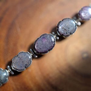 May include: A bracelet featuring a series of oval-shaped, dark reddish-purple cabochon stones set in scalloped metal bezels. The stones exhibit a natural, textured surface with visible inclusions, linked together by small metal connectors.