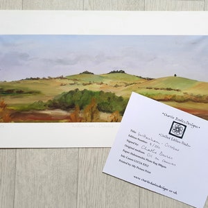 May include: A landscape painting titled "Wittenham - October" depicts rolling green hills under a cloudy sky. The artwork, signed by Charlie Davies, is an oil on canvas print, with a limited edition number of 8/250. A card with details is next to the artwork.