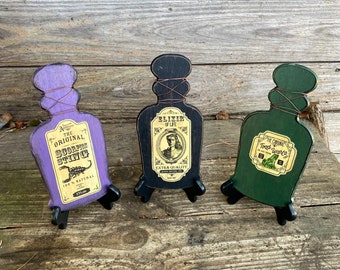 May include: Three wooden potion bottles with cork stoppers and wire accents. The bottles are painted in purple, black, and green. Each bottle has a label with a different potion name and illustration. The labels read "The Original Scorpion Sting", "Elixir of Life", and "The Original Toad Warts".