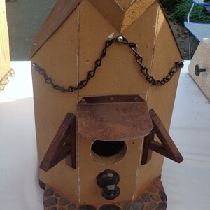 May include: A brown wooden birdhouse with a rusty metal roof and a chain hanging from the top. The birdhouse has a small opening for birds to enter and a stone base.