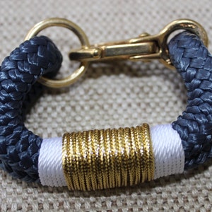 May include: A navy blue rope bracelet with a gold and white accent. The bracelet has a gold clasp.