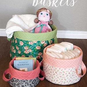 May include: Three fabric storage baskets with quilted rims. The largest is green with pink flowers, holding a doll and white blanket. A smaller pink basket holds diapers. The smallest is grey with floral patterns, containing a book titled The Tale of Tom Kitten.
