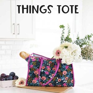 May include: A floral tote bag with pink trim and handles, filled with flowers and a baguette. The bag has a dark background with pink and orange flowers. The text "ALL THE THINGS TOTE" is above the bag.