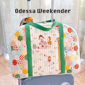 May include: A quilted "Odessa Weekender" travel bag with a green handle and zipper. The bag features a patchwork design with colorful hexagonal patterns and a central panel with a picnic scene illustration. The bag is on top of a blue suitcase.