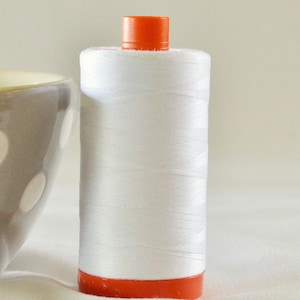 May include: A spool of white thread with an orange top and bottom. The thread is tightly wound around the spool, and the orange parts are a bright, solid color. The image is a close-up, showing the texture of the thread.