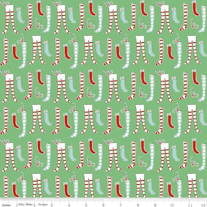 May include: A green fabric with a repeating pattern of red and white striped stockings and blue and white striped stockings. The stockings are arranged in a vertical pattern with a small green sprig of holly between each pair.