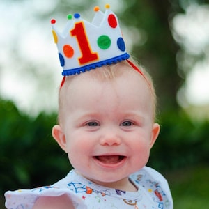 May include: A baby wearing a white felt crown with red, blue, green, and yellow polka dots and the number "1" in red felt. The crown has a blue felt trim and is decorated with white pom-poms.