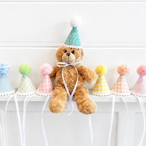 Pode incluir: Um urso de peluche castanho usa um chap&eacute;u de festa azul-petr&oacute;leo com um pompom branco e fitas brancas, rodeado por seis chap&eacute;us de festa em miniatura com padr&otilde;es axadrezados pastel, cada um com um pompom a condizer e rebordo branco.