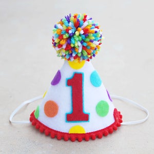 Puede incluir: Un gorro de fiesta de fieltro blanco con un n&uacute;mero uno rojo, lunares de colores y un pomp&oacute;n arco&iacute;ris en la parte superior.