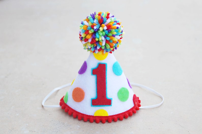 Puede incluir: Un gorro de fiesta de fieltro blanco con un n&uacute;mero uno rojo, lunares de colores y un pomp&oacute;n arco&iacute;ris en la parte superior.