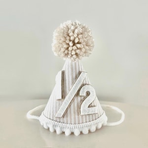 Puede incluir: Un gorro de fiesta a rayas beige y blanco, adornado con el n&uacute;mero "1/2" en blanco. Tiene un pomp&oacute;n esponjoso en la parte superior y un ribete de pomp&oacute;n blanco. Una cuerda blanca est&aacute; unida para asegurar el gorro. Ideal para un medio cumplea&ntilde;os.