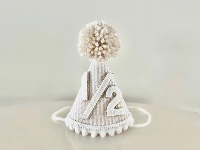 Puede incluir: Un gorro de fiesta a rayas beige y blanco, adornado con el n&uacute;mero "1/2" en blanco. Tiene un pomp&oacute;n esponjoso en la parte superior y un ribete de pomp&oacute;n blanco. Una cuerda blanca est&aacute; unida para asegurar el gorro. Ideal para un medio cumplea&ntilde;os.