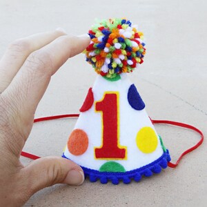 May include: A white felt party hat with a red felt number one, colorful polka dots, and a blue pom-pom trim. The hat has a red elastic band.