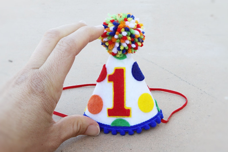 May include: A white felt party hat with a red felt number one, colorful polka dots, and a blue pom-pom trim. The hat has a red elastic band.