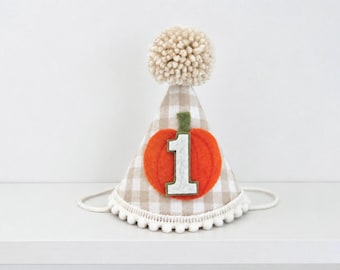Op de afbeelding: Een kegelvormige feesthoed met een beige en wit ruitpatroon, bekroond met een grote beige wollen pompon. De onderrand is afgewerkt met kleine witte pompons. Centraal bevindt zich een oranje viltapplicatie in de vorm van een pompoen met een groen steeltje.