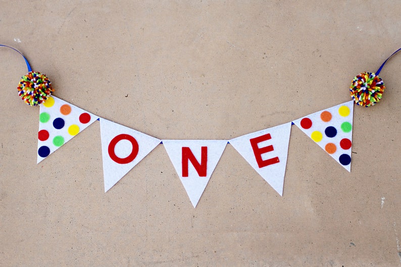 May include: A white felt banner with the word "ONE" in red letters. The banner is decorated with colorful polka dots and has blue ribbon ties on each end.