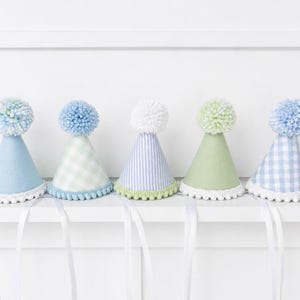 Puede incluir: Cinco gorros de fiesta cónicos en tonos pastel, que incluyen azul claro liso, cuadros vichy azul claro, rayas azul claro, verde salvia liso y cuadros vichy azul claro. Cada gorro tiene un pompón de lana a juego y un borde blanco.