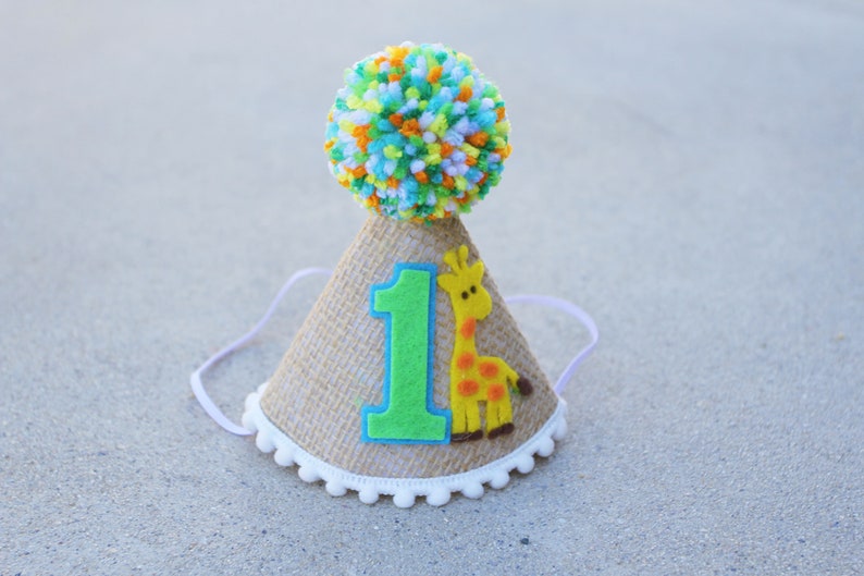 May include: A brown burlap party hat with a white pom-pom on top. The hat has a green felt number "1" and a yellow felt giraffe with orange spots.