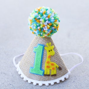 May include: A brown burlap party hat with a white pom-pom on top. The hat has a green felt number one and a yellow felt giraffe with orange spots.