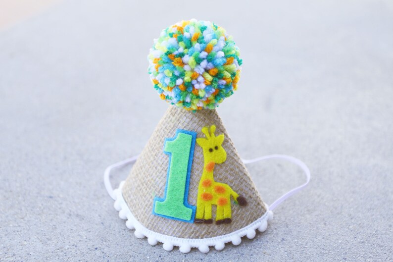 May include: A brown burlap party hat with a white pom-pom on top. The hat has a green felt number one and a yellow felt giraffe with orange spots.