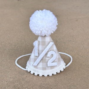 May include: A beige and white gingham party hat with a white pom-pom on top. The hat has a white felt applique with the number "1/2" on it.