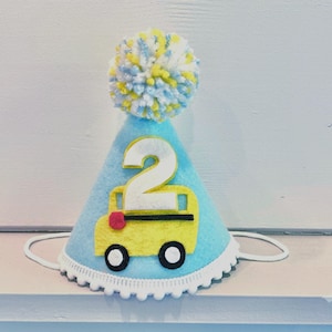 May include: A light blue felt birthday party hat featuring a yellow school bus and the number 2. The hat has a white scalloped trim and a pom-pom in shades of yellow and blue. A white elastic band is attached.