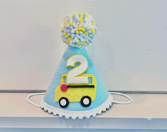 Op de afbeelding: Lichtblauwe vilten verjaardagshoed met een gele schoolbus en het nummer 2. De hoed heeft een witte geschulpte rand en een pompon met blauw en geel garen. Er is een wit elastisch touwtje bevestigd.
