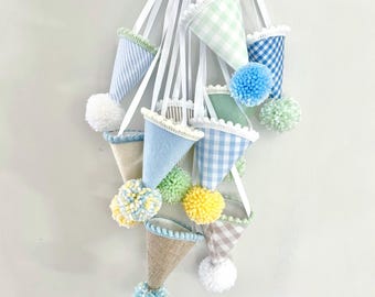 Pode incluir: Uma coleção de ornamentos suspensos em forma de cone com vários padrões de tecido, incluindo xadrez vichy, riscas e cores sólidas. Cada cone é rematado com uma fita branca e um pompom decorativo em tons de azul, amarelo, azul ou branco.