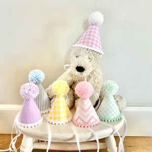 May include: A collection of six cone-shaped party hats featuring various pastel colors and patterns including gingham, stripes, and chevrons. Each hat is topped with a fluffy pom-pom and finished with a white pom-pom trim along the bottom edge.