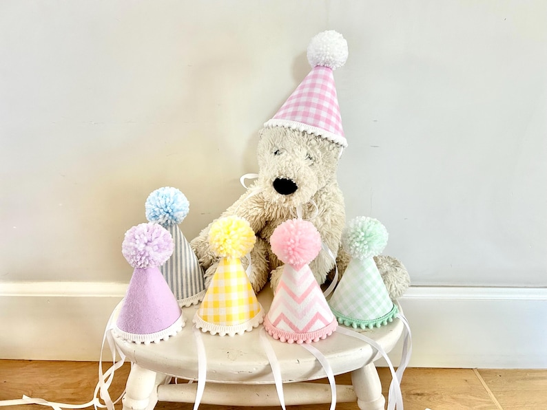 May include: A collection of six cone-shaped party hats featuring various pastel colors and patterns including gingham, stripes, and chevrons. Each hat is topped with a fluffy pom-pom and finished with a white pom-pom trim along the bottom edge.