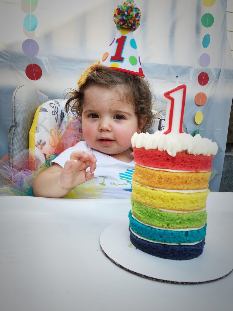 Puede incluir: Un ni&ntilde;o peque&ntilde;o con un sombrero de cumplea&ntilde;os colorido se sienta en una silla alta con un pastel de capas de arco&iacute;ris frente a &eacute;l. El pastel est&aacute; decorado con glaseado blanco y una vela con el n&uacute;mero uno.