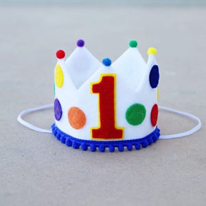 May include: White felt crown with a red felt number one and colorful felt dots. The crown has a blue felt trim and a white elastic band.