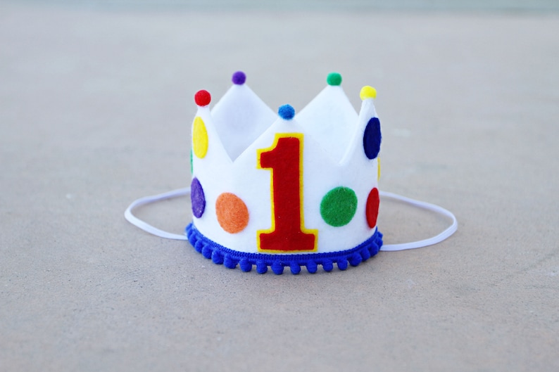 May include: White felt crown with a red felt number one and colorful felt dots. The crown has a blue felt trim and a white elastic band.