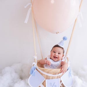 Puede incluir: Un beb&eacute; sonr&iacute;e desde una cesta de mimbre, parte de un montaje de globo aerost&aacute;tico. El globo es de color rosa claro. La cesta tiene detalles azules y blancos, y el beb&eacute; lleva un gorro y un babero a juego.