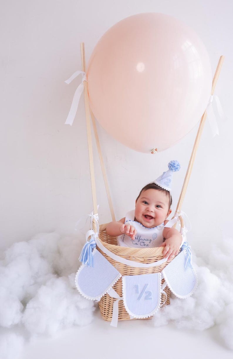Puede incluir: Un beb&eacute; sonr&iacute;e desde una cesta de mimbre, parte de un montaje de globo aerost&aacute;tico. El globo es de color rosa claro. La cesta tiene detalles azules y blancos, y el beb&eacute; lleva un gorro y un babero a juego.