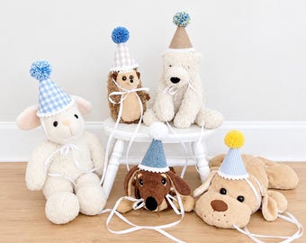 May include: A collection of five plush animals including a lamb, hedgehog, polar bear, dachshund, and dog, each wearing a miniature party hat. The hats feature various patterns like blue gingham, stripes, and solid colors, topped with colorful yarn pom-poms.