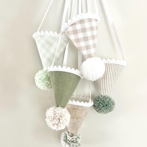 Puede incluir: Un móvil colgante con seis adornos en forma de cono hechos de diversas telas, incluyendo fieltro verde salvia, cuadros vichy beige y patrones de rayas. Cada cono tiene un borde de pompones blancos y un pompón de lana colgando.