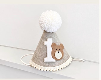 May include: A cone-shaped party hat made of light gray felt, topped with a large white yarn pom-pom. The front features a white felt number one and a brown bear face appliqué. The bottom edge is trimmed with a white pom-pom fringe.