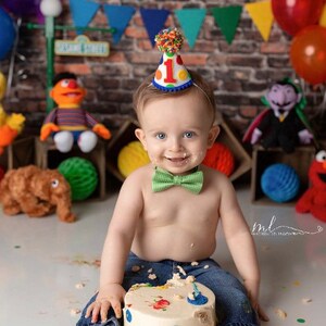 May include: A baby boy wearing a blue and red birthday hat with a number one on it, a green bow tie, and blue jeans, is sitting in front of a birthday cake with a Sesame Street theme. The cake is decorated with colorful sprinkles and a Sesame Street character.