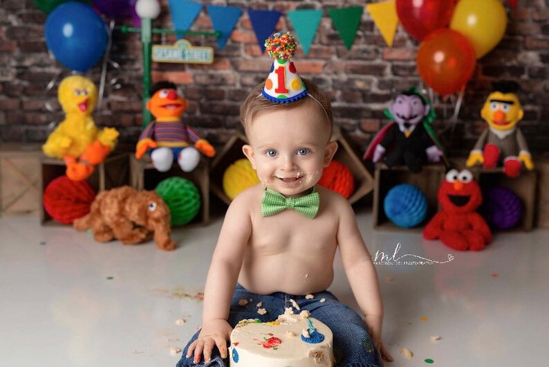 May include: A baby boy wearing a blue and red birthday hat with a number one on it, a green bow tie, and blue jeans, is sitting in front of a birthday cake with a Sesame Street theme. The cake is decorated with colorful sprinkles and a Sesame Street character.