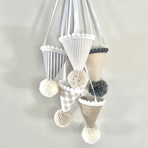 May include: A hanging mobile featuring five cone-shaped ornaments suspended by white ribbons. The cones display various patterns including grey stripes, beige gingham, and solid neutral tones, each finished with a pom-pom and white trim.