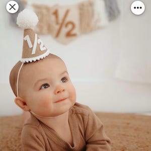 Puede incluir: Un beb&eacute; con un gorro de fiesta de arpillera con el texto "1/2" en blanco. El gorro est&aacute; adornado con un pomp&oacute;n blanco y un ribete festoneado blanco. El beb&eacute; lleva un su&eacute;ter marr&oacute;n y est&aacute; tumbado sobre una alfombra marr&oacute;n.