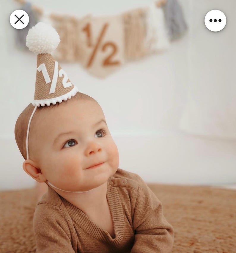 Puede incluir: Un beb&eacute; con un gorro de fiesta de arpillera con el texto "1/2" en blanco. El gorro est&aacute; adornado con un pomp&oacute;n blanco y un ribete festoneado blanco. El beb&eacute; lleva un su&eacute;ter marr&oacute;n y est&aacute; tumbado sobre una alfombra marr&oacute;n.