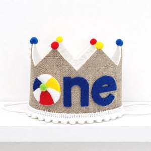May include: A tan burlap crown featuring a white, red, yellow, and blue beach ball applique next to the word one in blue felt letters. The crown is topped with small red, yellow, and blue pom-poms and has a white pom-pom trim along the bottom edge.