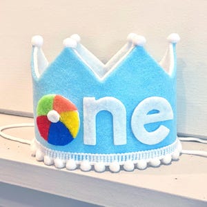 Puede incluir: Una corona de fieltro azul claro con una aplicación de pelota de playa colorida y la palabra one en letras de fieltro blancas. La corona está decorada con un borde de pompones blancos en la base y en las puntas, con lazos blancos.