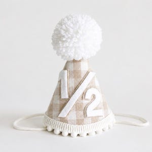 May include: A cone-shaped party hat featuring a beige and white gingham pattern. The front displays the white felt fraction 1/2. The hat is topped with a large white yarn pom-pom and finished with a white pom-pom trim along the bottom edge.