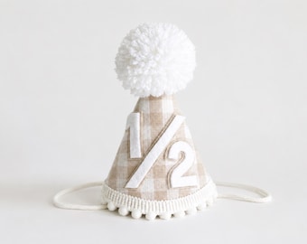 May include: A cone-shaped party hat featuring a beige and white gingham pattern. The front displays the white felt fraction 1/2. The hat is topped with a large white yarn pom-pom and finished with a white pom-pom trim along the bottom edge.