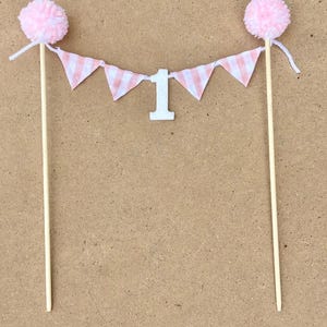 May include: Pink and white gingham banner with the number "1" in the center, attached to two wooden sticks with pink pom poms on top.