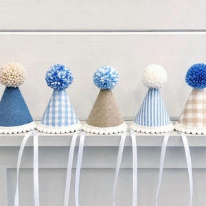 Pode incluir: Cinco chap&eacute;us de festa em forma de cone com acabamento de pompom branco na base e um grande pompom de l&atilde; no topo. Os chap&eacute;us apresentam v&aacute;rios padr&otilde;es, incluindo ganga azul s&oacute;lida, xadrez vichy azul e branco, linho bege, riscas.