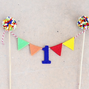 May include: A festive cake topper featuring two pom-poms with colorful yarn and a banner of felt pennants in green, orange, red, and yellow. A blue number 1 is centered on the banner, perfect for a first birthday celebration.